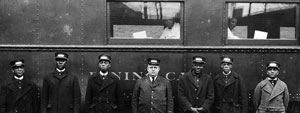 A group of black porters and a white man are standing on the platform next to the dining car. Two black men can be seen in the window.